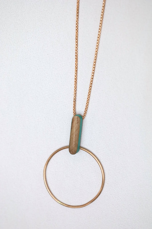 Bronze circle and wooden dash pendant suspended from bronze chain necklace, on a light background.