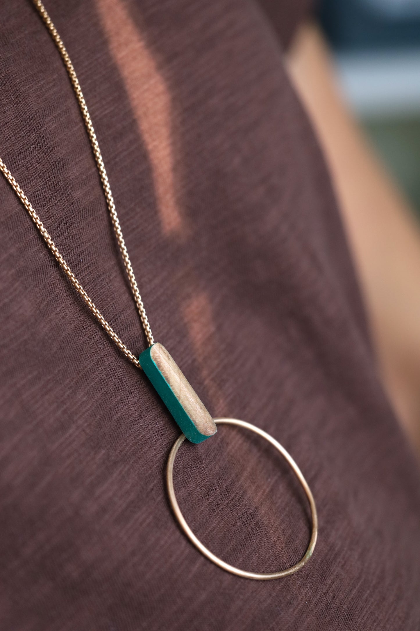 Bronze circle and wooden dash pendant suspended from bronze chain necklace. Side angle on dark background.