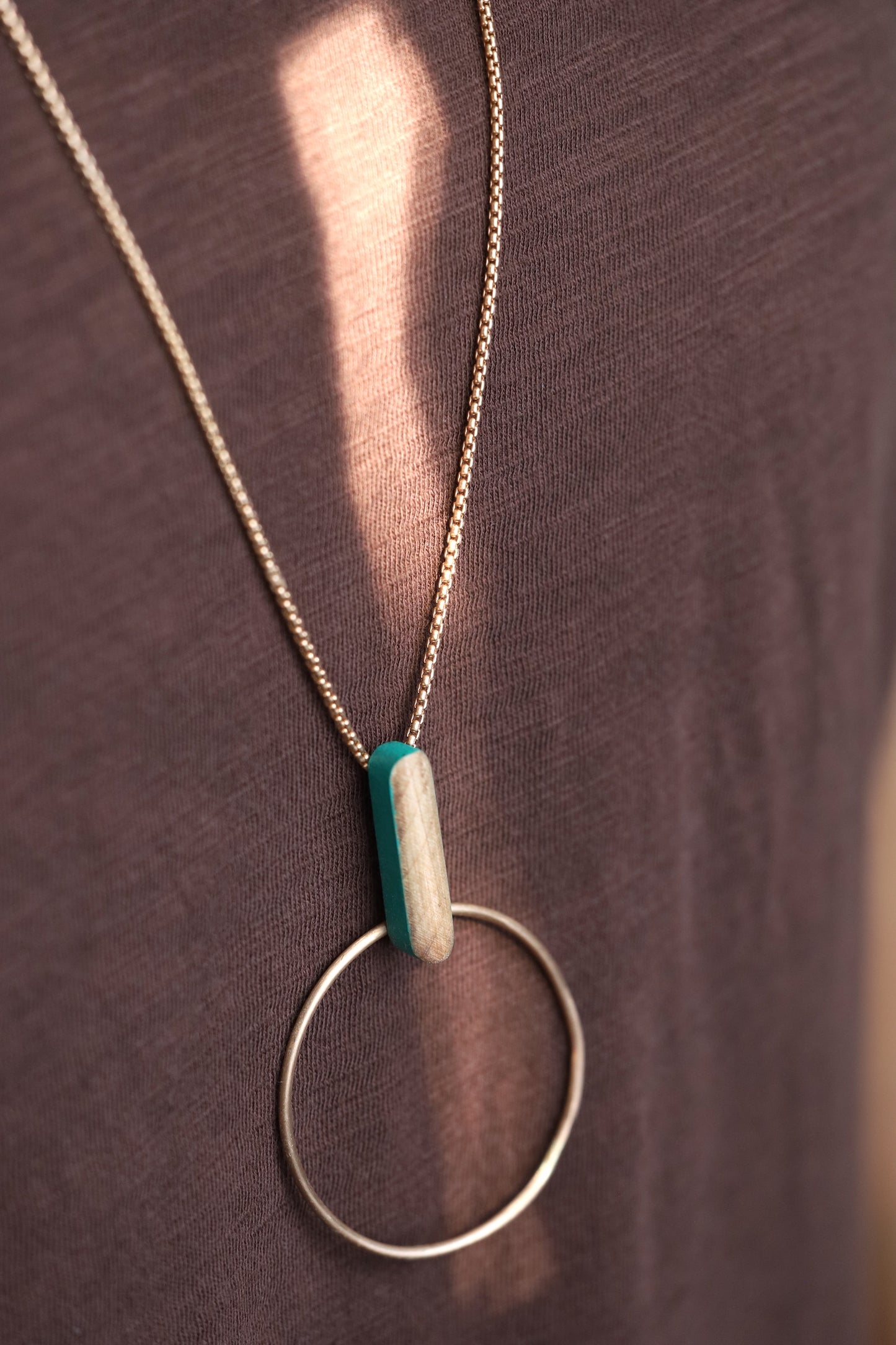 Bronze circle and wooden dash pendant suspended from bronze chain necklace. Slight angle on dark background.