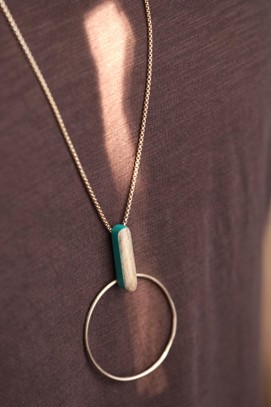 Bronze circle and wooden dash pendant suspended from bronze chain necklace. Slight angle on dark background.