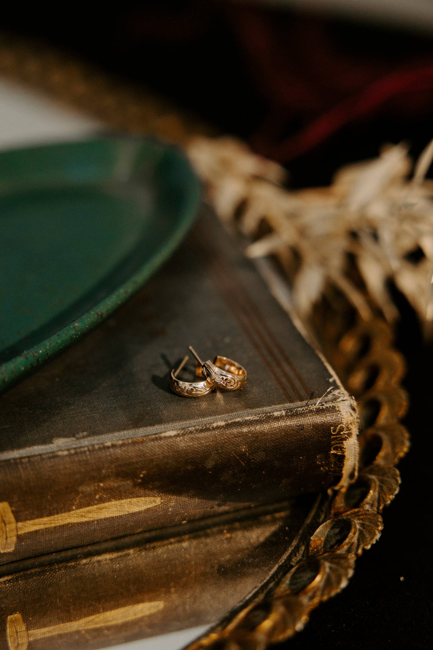A pair of gold-toned textured hoop earrings resting on a vintage dark brown book, styled with a green plate and dried grass on a dark background.