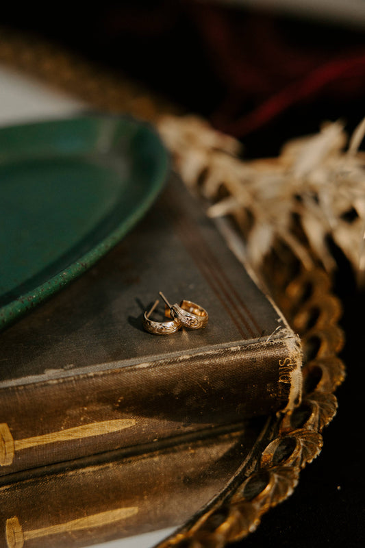A pair of gold-toned textured hoop earrings resting on a vintage dark brown book, styled with a green plate and dried grass on a dark background.