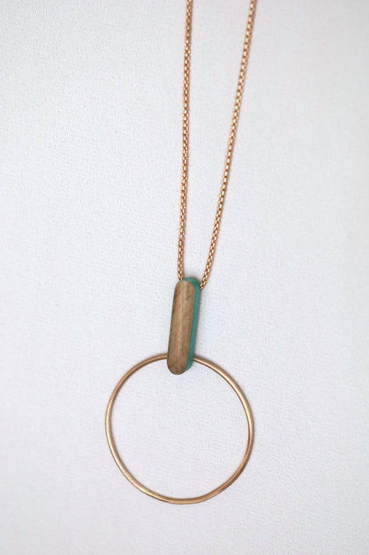 Bronze circle and wooden dash pendant suspended from bronze chain necklace, on a light background.