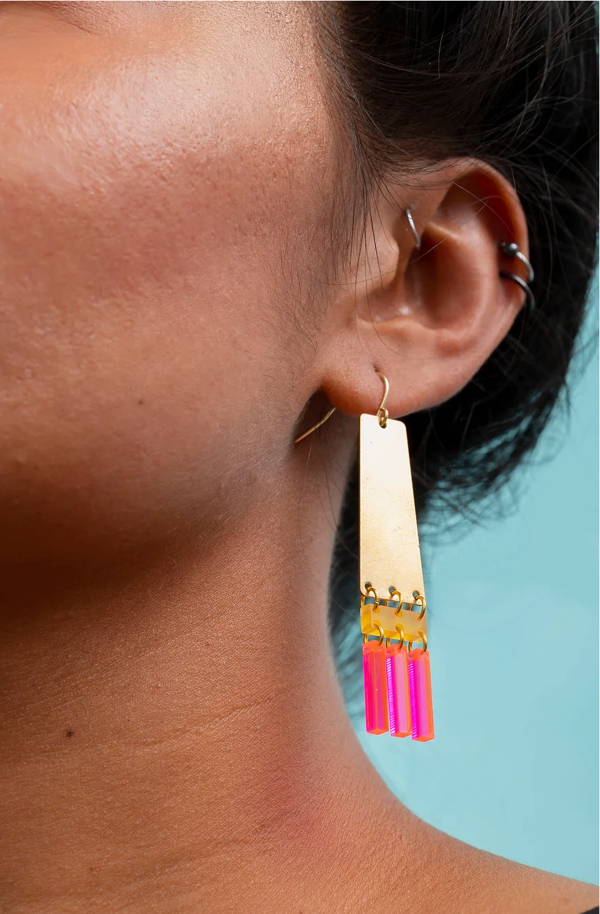 Woman wearing brass and colorful acrylic earrings with pink fringe.